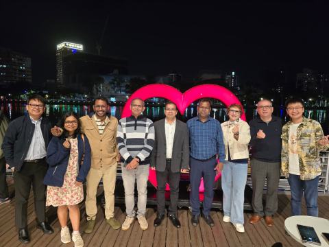 Speakers and hosts pose for a group photo at night in front of a large illuminated red heart on the riverside, with city lights reflected in the water.