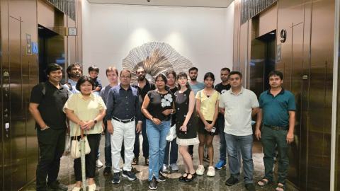 Group photo of lab members, family members and a child in an elevator lobby, in front of a silver sculpture on the wall.
