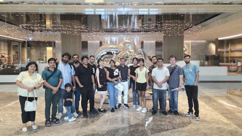 Group photo of lab members, family members and a child in a large lobby, in front of a chrome sculpture under a sparkling ceiling installation.