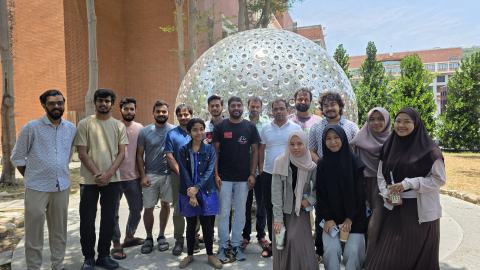 Group photo of lab members outdoors in front of a large silver sphere sculpture and a red-brick building.